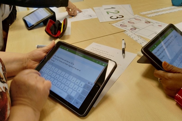Adults are learning Swedish. The learners sit at a table facing each other, and each is holding a tablet in their hands. The learners are writing descriptions of a person in Swedish. On the table, there are handouts, a ballpoint pen and a spectacle case.