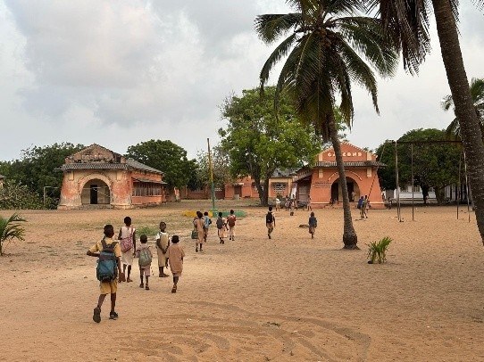 Koululaisia kävelee kohti koulurakennuksia hiekkapihalla Beninissä, Länsi-Afrikassa. Koululaisilla on reput selässään.